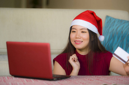 年轻美丽和快乐的亚洲亚洲妇女在圣诞节圣诞老人帽子拿着信用卡使用笔记本电脑上网购物微笑着兴奋在家里的沙发客厅