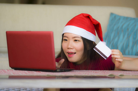 年轻美丽和快乐的亚洲亚洲妇女在圣诞节圣诞老人帽子拿着信用卡使用笔记本电脑上网购物微笑着兴奋在家里的沙发客厅