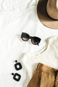 时尚柔和的构图与女装和配饰。米色长裤衬衫帽子太阳镜白色毯子上的耳环。平面布局, 顶部视图