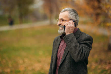 一个英俊的戴眼镜胡子的老人正在用电话。秋天在公园散步