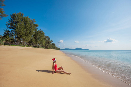 穿着红色泳衣和圣诞老人帽的年轻女子坐在宽阔的热带海滩上