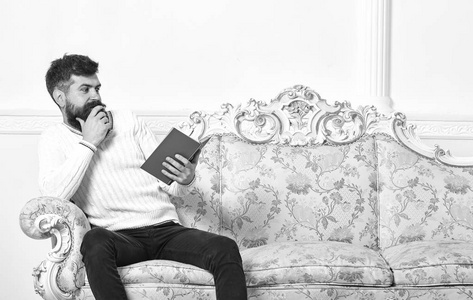 可耻的畅销书概念。男孩惊奇地读书。男子胡子和胡子坐在巴洛克风格的沙发上, 持有书籍, 白色的墙壁背景。男子气概惊讶的脸阅读书