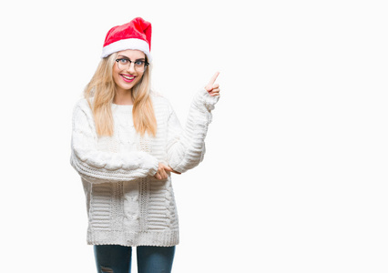 年轻美丽的金发女子戴着圣诞节帽子在孤立的背景与一个大微笑在脸上, 指着手和手指在一边看着相机