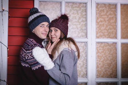年轻夫妇在毛衣和帽子一起在红墙附近的小房子装饰为玛丽圣诞节和新年