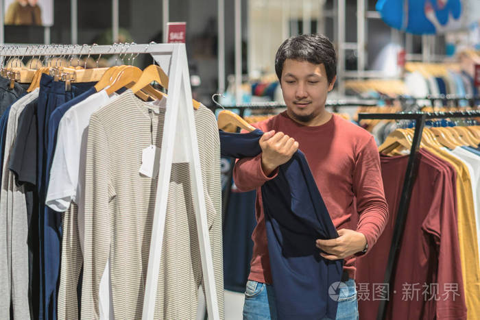 聪明的亚洲男人与胡子选择衣服在商店在购物中心, 看着五颜六色的 t恤