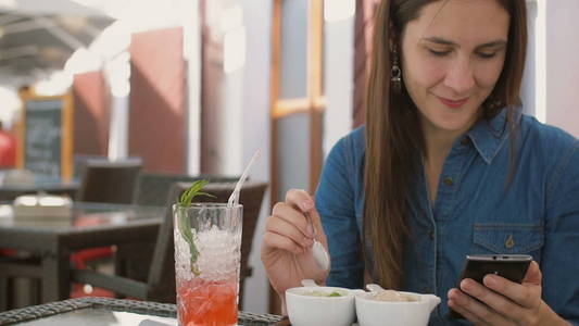 黑发的女孩坐在外面的一家咖啡馆，同时使用了智能手机，吃冰激淋。4 k