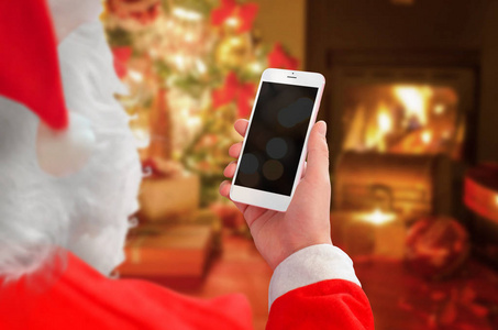 圣诞老人举行白色智能手机样机的空白屏幕。圣诞节的时候带壁炉的背景