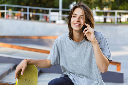 漂亮的年轻滑板手坐在公园里, 用手机说话的滑板的图像
