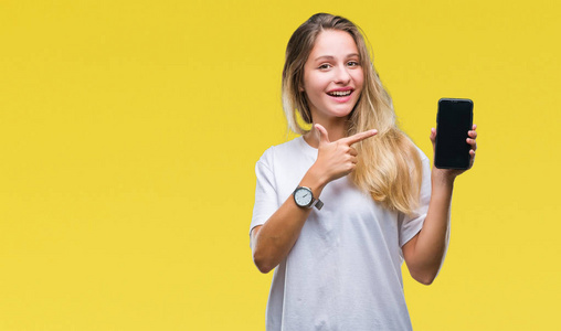 年轻美丽的金发女子显示屏幕的智能手机在孤立的背景非常愉快的指着手和手指