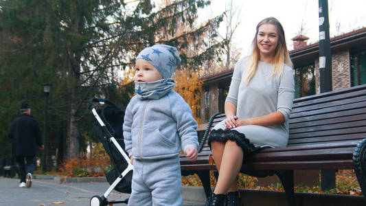 年轻的母亲坐在长凳上, 她的小宝宝走在公园里