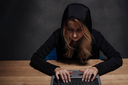 专注的女性黑客在黑色连帽使用笔记本电脑在木桌面