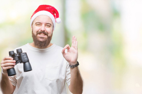 年轻的高加索嬉皮士男子戴着圣诞帽, 看起来虽然双筒望远镜在孤立的背景做确定的标志与手指, 优秀的象征