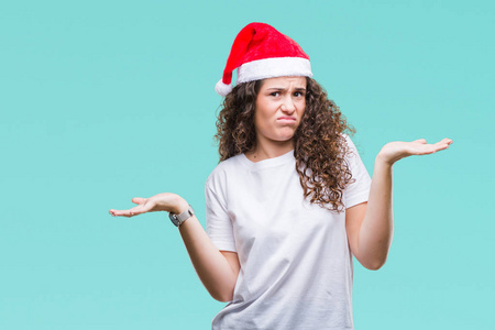 年轻的黑发女孩戴着圣诞帽在孤立的背景无线索和困惑的表情与手臂和手抬起来。怀疑概念