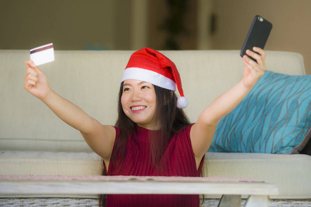 年轻快乐和美丽的亚洲亚洲妇女在圣诞老人帽子持有信用卡使用手机圣诞网上购物从家里的沙发微笑着愉快的互联网商业概念