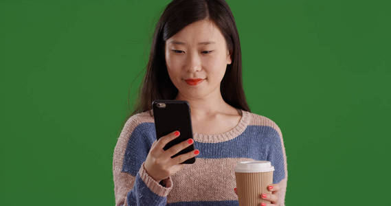 亚洲妇女在使用手机在绿色屏幕上发短信时喝咖啡的肖像