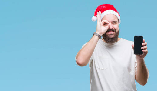 年轻的高加索男子戴着圣诞帽, 显示智能手机屏幕在孤立的背景与快乐的脸微笑做确定的标志与眼睛看着通过手指