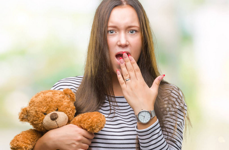 年轻的高加索妇女抱着泰迪熊在孤立的背景覆盖嘴与手震惊与耻辱, 表达恐惧, 害怕在沉默中, 秘密的概念