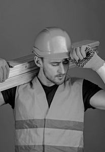 木匠, 木工, 强的建设者在繁忙的面孔运载木横梁在肩膀上。安全和保护概念。戴防护手套的男子持有防护硬帽子遮阳板, 灰色背景