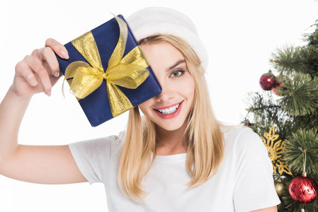 微笑的妇女在圣诞老人帽子持有包裹的礼物附近的圣诞树查出的白色