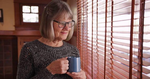 甜蜜的老年妇女与咖啡杯看窗外从客厅