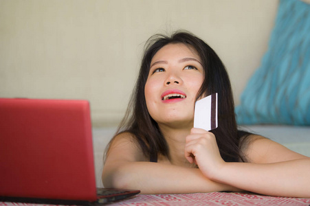 年轻美丽和快乐的亚洲韩国妇女持有信用卡银行和网上购物与笔记本电脑在家里的沙发在互联网业务和电子商务概念的生活方式肖像