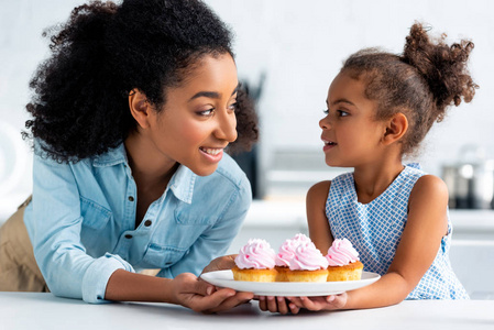 微笑的非洲裔美国母亲和女儿拿着自制的纸杯蛋糕, 看着对方在厨房