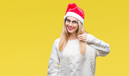 年轻美丽的金发女子戴着圣诞节帽子在孤立的背景做愉快的拇指手势与手。批准表达式看着相机显示成功