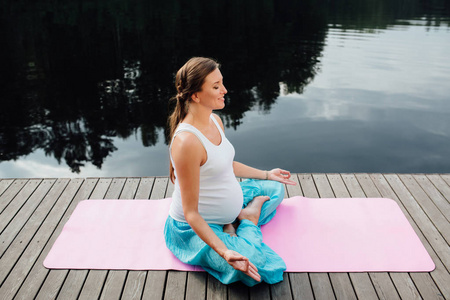 年轻女子在荷花的立场练瑜伽在河边森林。坐在垫子上木码头