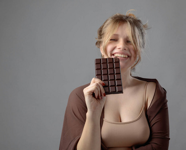 快乐的年轻美丽的女人吃巧克力。年轻的金发碧眼与自然化妆有乐趣和吃巧克力孤立的灰色背景