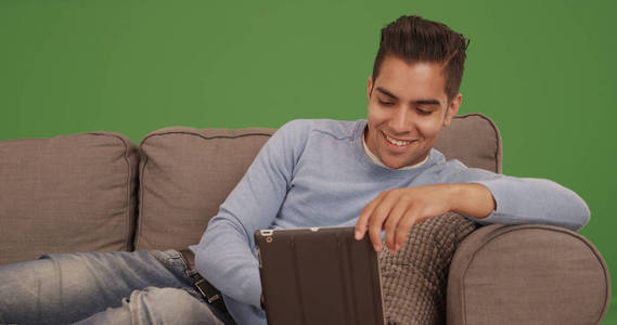 年轻的西班牙男子坐在沙发上使用平板电脑在绿色屏幕上