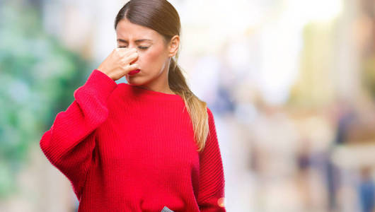 年轻美丽的商业妇女穿着冬季毛衣在孤立的背景闻到一些臭和恶心, 无法忍受的气味, 用手指在鼻子上屏住呼吸。难闻的气味概念