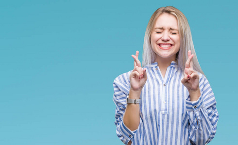 年轻的金发女人在孤立的背景下微笑着交叉手指, 希望和眼睛闭上。运气和迷信概念