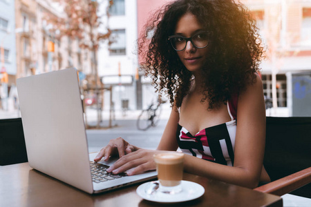 迷人的高加索年轻女性自由职业者在眼镜和卷曲的笨重的头发是在一个户外咖啡馆的笔记本电脑上工作与一杯美味的咖啡旁边, 她在桌子上