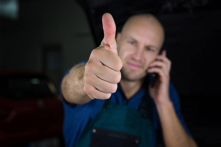 年轻的机修工在一家汽车商店里展示了一个隆起的拇指, 而他打电话