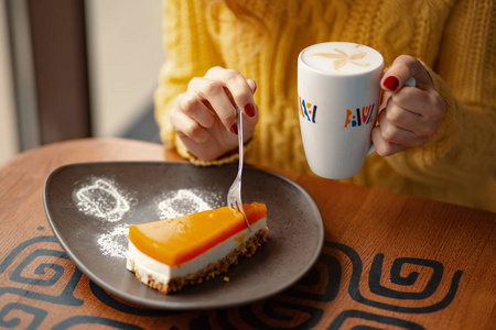 在一张木桌上, 妇女手上的一小片美味的胡萝卜芝士蛋糕和拿铁咖啡杯。顶部角度视图