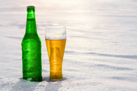 杯和瓶冰镇啤酒，在日落时雪。美丽的冬天背景。户外娱乐。含酒精饮料的广告