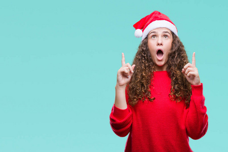 年轻的黑发女孩在孤立的背景下戴着圣诞帽, 惊讶和惊讶地抬头, 用手指指指点点, 举起双臂