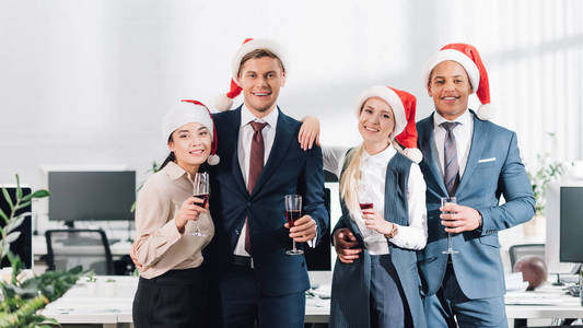 快乐的年轻同事在圣诞老人帽子喝酒, 微笑着在镜头前, 而庆祝新年在办公室