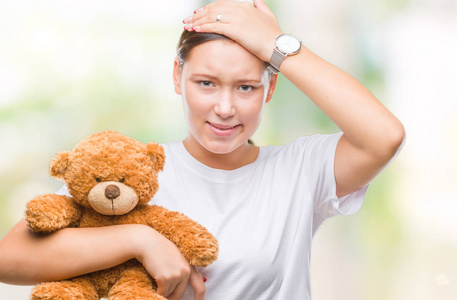 年轻的高加索妇女抱着泰迪熊在孤立的背景上用手在头上强调, 震惊与羞愧和惊讶的脸, 愤怒和沮丧。因错误而恐惧和不安