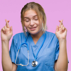 年轻的高加索医生妇女穿着外科医生制服在孤立的背景微笑地交叉手指与希望和眼睛闭合。运气和迷信的概念