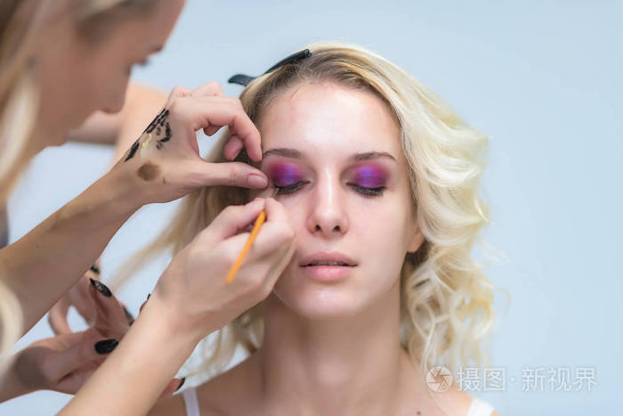 一个专业化妆师的作品美容师, 化妆与画笔在一个美丽的金发女郎的脸上与阴影的模型的眼睛。眉毛的绘画