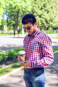 一名男子站在城市里，端着咖啡或者茶，在夏天在眼镜和一件衬衫在公园户外休息商人读取通电话，社交网络上的消息