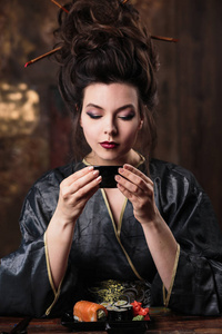 穿着艺妓亚洲服装的感性年轻女子, 穿着时尚化妆和发型的服装, 喝着茶, 吃寿司, 时装美像
