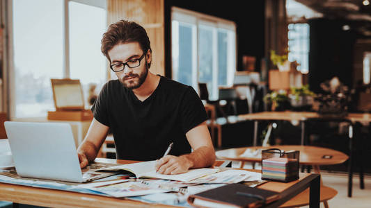 专注于工作项目的平面设计师。年轻的胡子画师的肖像坐在工作场所看在笔记本电脑屏幕写在记事本室内。创意工作理念