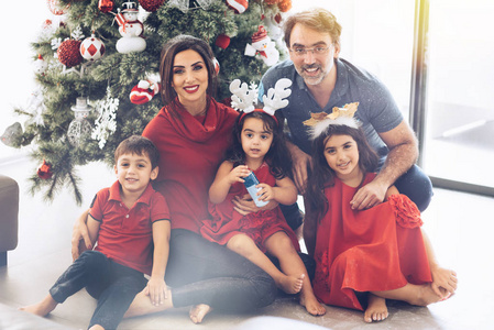 圣诞节和新年主题的父母和孩子坐在家里的圣诞树下拥抱着看着的相机, 微笑着愉快地在一起