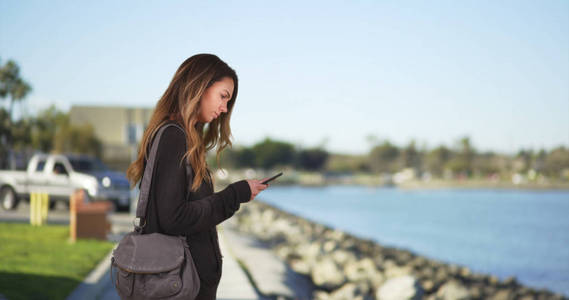 年轻的千禧女孩在海湾边的手机上发短信微笑和大笑