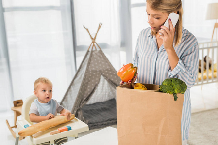 忙碌的母亲拿着杂货, 而在智能手机上交谈与幼儿在婴儿椅子上的背景