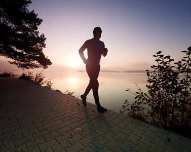 穿着时髦跑鞋和运动服的高个子跑步运动员在岸边晨练的冻结动作。路面轨道在湖附近