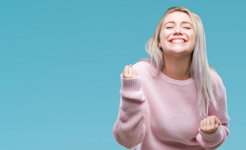 年轻的金发女人穿着冬季毛衣在孤立的背景下非常高兴和兴奋做赢家手势与手臂举起, 微笑和尖叫的成功。庆祝概念