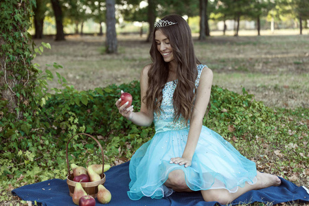 美丽的年轻女孩头上戴着皇冠, 穿着蓝色的衣服, 坐在草地上的毯子上。她很高兴, 面带微笑。她手里拿着一个苹果。在她旁边有一个篮子